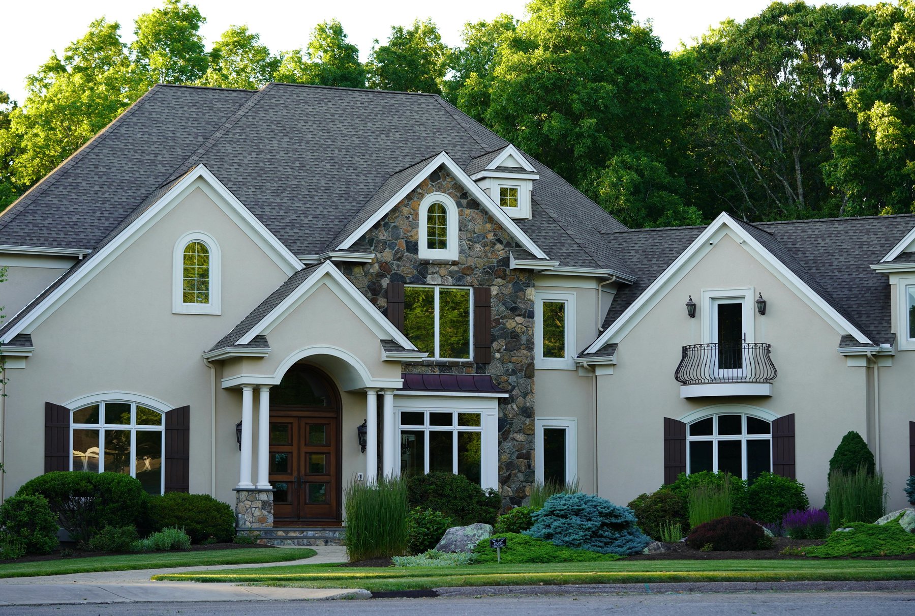 A two-story cream stucco home with a stone entry tower, dark wood doors, white trim, and a manicured front lawn framed by tall trees.