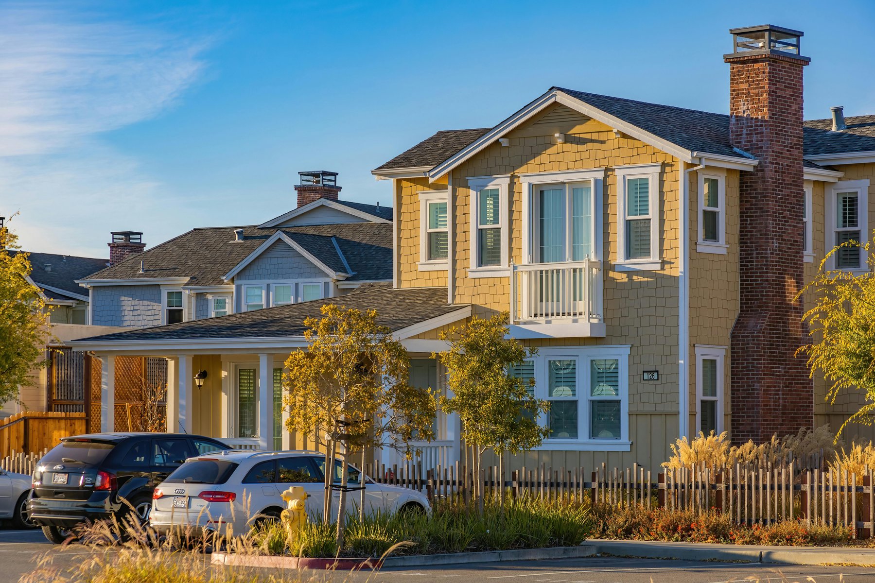 A yellow two-story house and a light blue neighbor behind a small parking lot with several cars, a yellow fire hydrant, and a clear blue sky.