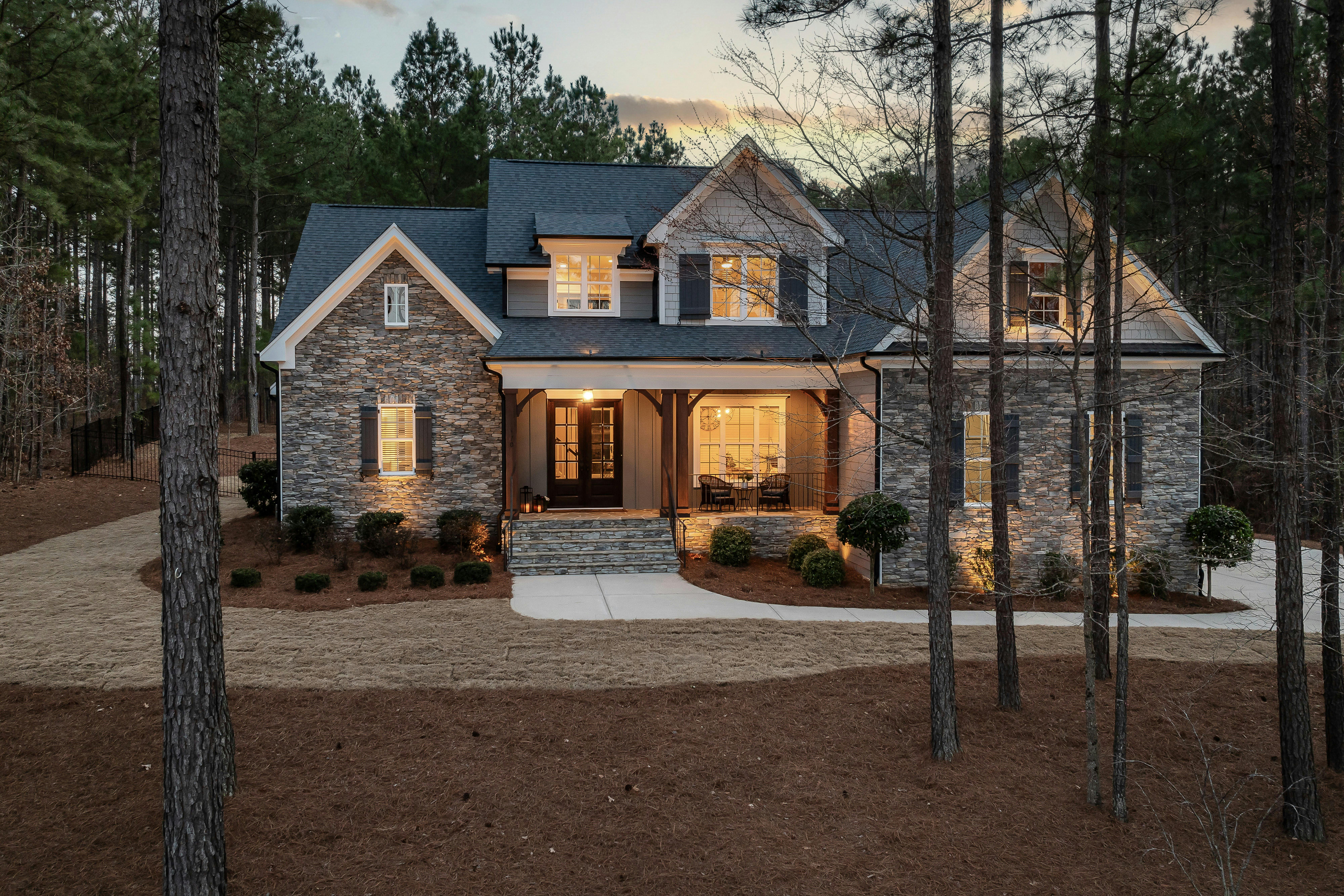 A two-story stone and siding house at dusk, set back among tall pines with a lit front porch, a curved concrete walk, and a long drive across a pine straw lawn.